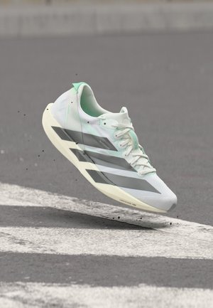 Chaussure de course blanche et vert menthe avec des rayures noires, en l'air au-dessus d'une surface en asphalte noir avec des marquages routiers blancs.