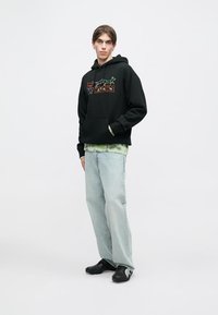 Melnā kapucēta džemperis ar priekšējo grafiku, ar kabatu priekšā; virsū uz zaļās tīrās krāsas krekla. Gaiši zili platie džinsi un melna apavi pabeidz izskatu.