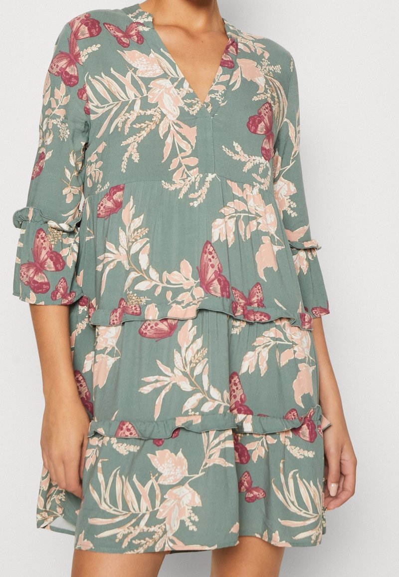 Robe verte à fleurs avec des papillons roses, jupe à étages, manches 3/4 avec poignets noués et encolure en V portée par une personne les mains sur les côtés.