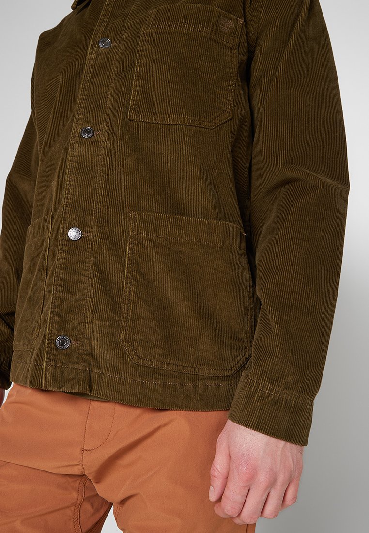 Brązowa kurtka z korduroy z zatrzaskami, dwiema przednimi kieszeniami, teksturowanym materiałem i luźnym krojem, połączona z pomarańczowymi spodniami.