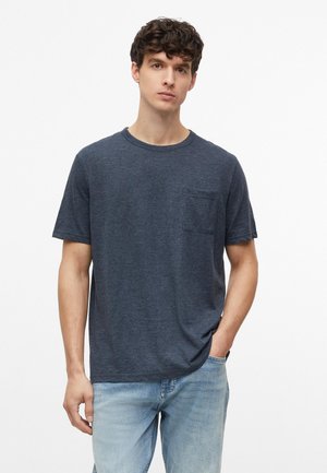 Junger Mann mit lockigem Haar, der ein dunkelgraues kurzärmeliges T-Shirt mit Brusttasche und hellblaue Jeans trägt, steht mit einer Hand in der Tasche.