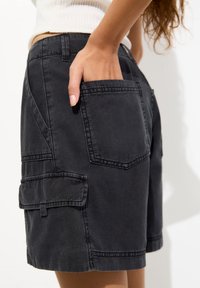 Bermudas cargo de mezclilla negra con múltiples bolsillos, detalles de costura y un corte relajado. La tela parece suave con una textura ligeramente desgastada.