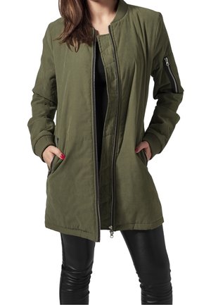 Mujer con una chaqueta bomber verde oliva con bolsillos con cremallera y pantalones de cuero negro, con las manos en los bolsillos de la chaqueta, de pie frente a un fondo blanco.