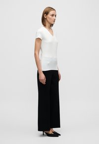 Λευκό τοπ με κοντά μανίκια και V-λαιμόκοψη, συνδυασμένο με μαύρο παντελόνι φαρδιάς γραμμής και μαύρες γόβες. Λείο ύφασμα, εφαρμοστό στο σώμα, ρέον στην περιοχή των γοφών.