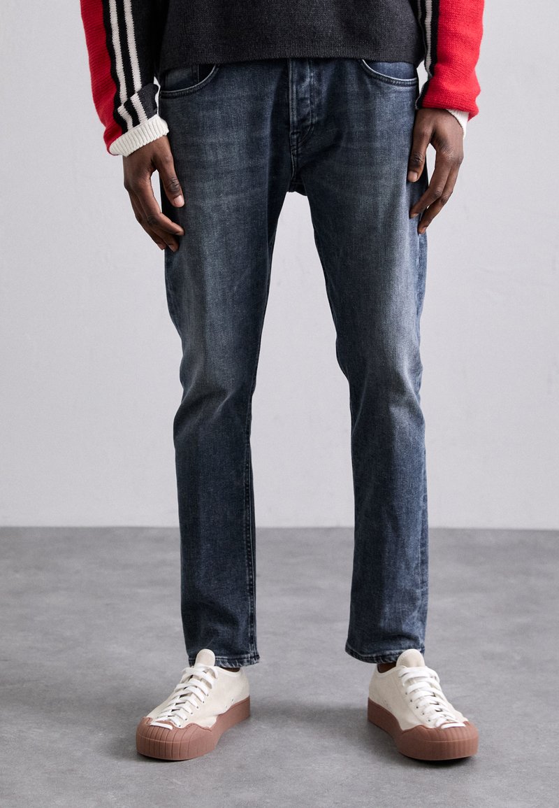 Jeans in denim blu scuro con taglio dritto, abbinati a sneakers off-white con suola in gomma marrone testurizzata.