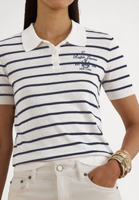 Sieviete valkā baltu un tumši zilu svītrainu polo krekla ar izšūtu logo un baltas bikses, papildinātas ar vairākiem zelta rokassprādzēm.