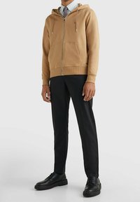 Beige hoodie met rits, voorzakken, ribgebreide manchetten en een capuchon met trekkoord, gecombineerd met zwarte broeken en zwarte schoenen.