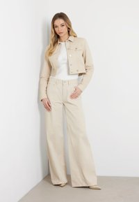 Giacca cropped beige con colletto arrotondato e hardware silver, abbinata a pantaloni beige a gamba larga. Sotto, una maglietta bianca a coste.