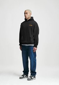 Black hoodie with pocket and orange logo, paired with blue denim jeans and black-and-white sneakers. Smooth fabric with a relaxed fit.
