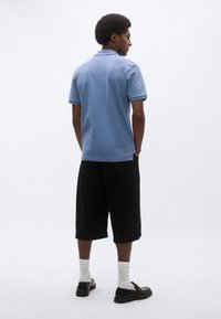 Άνδρας με αφροαμερικανικό κούρεμα, φοράει ανοιχτό γαλάζιο πόλο με κοντά μανίκια, μαύρες σορτς μέχρι το γόνατο, λευκές κάλτσες και μαύρα παπούτσια, στέκεται πλάγια.