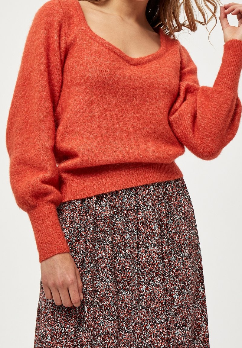 Kuscheliger, orangefarbener Strickpullover mit tiefem V-Ausschnitt und Puffärmeln, kombiniert mit einem schwarz-roten Rock mit floralen Mustern.