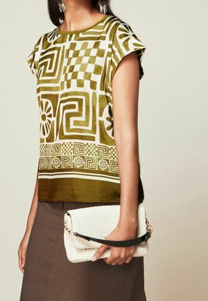 Frau trägt olivegrünes, gemustertes Kurzarmoberteil, braunen Rock, hält weiße, gewebte Clutch mit schwarzem Riemen.