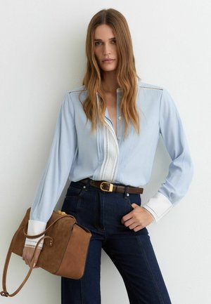 Femeie cu păr lung și castaniu, purtând o bluză bleu cu manșete albe, blugi închiși la culoare cu o curea maro, ținând o geantă maro din piele întoarsă.