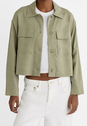 Personne portant une veste courte olive clair sur un crop top blanc côtelé et un pantalon blanc taille haute, debout devant un fond uni.