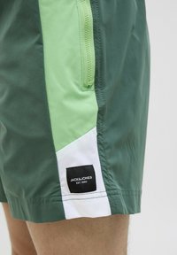 Groene en lichtgroene zwembroek met witte accenten, voorzien van een ritszak en een zwart label met witte tekst. Glad, lichtgewicht stof.