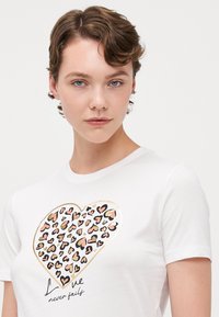 Witte t-shirt met korte mouwen, met een hartgrafiek in een luipaardprintpatroon in roze, zwart en gouden accenten, met tekst eronder.