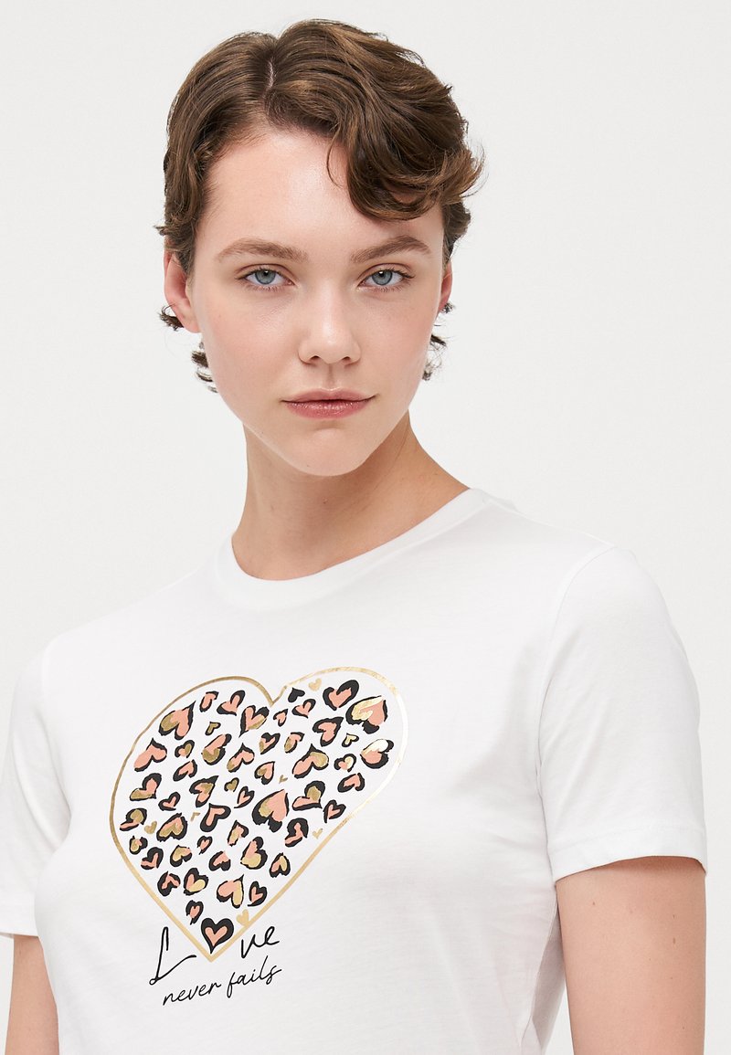 Camiseta blanca de manga corta con un gráfico de corazón en un patrón de leopardo en rosa, negro y detalles en dorado, con texto debajo.