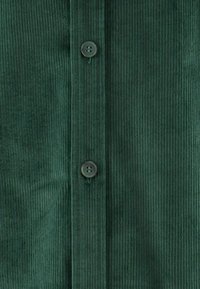 Donkergroene corduroy overhemden met verticale ribbelstructuur, twee ronde, grijze knopen en een schone, rechte snit langs de knoopsluiting.