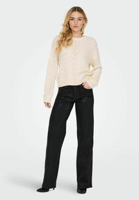 Pull en maille crème avec un motif en zigzag texturé et ourlet côtelé, associé à un pantalon noir large taille haute et des chaussures noires.