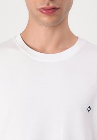 White cotton t-shirt with a round neckline, featuring a small black logo on the left chest area. Smooth texture, simple design.