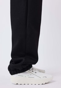 Zwarte sweatpants met elastische boorden, gecombineerd met witte sneakers met gestructureerde zolen en zwarte lijnaccenten, zijaanzicht.