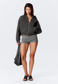 Gray cropped hoodie with zipper, paired with gray shorts. Holding a black bag with silver studs. Black slip-on sandals. Clean background.