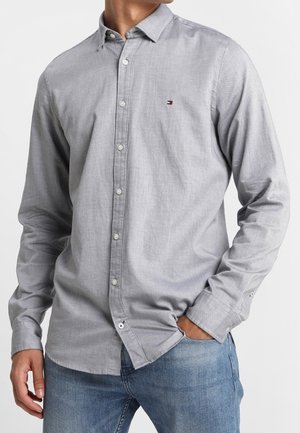 Homme portant une chemise gris clair boutonnée à manches longues et un jean bleu, une main dans la poche.