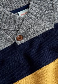 Nahaufnahme eines gestrickten Pullovers mit grauem Kragen, braunem Knopf, horizontalen Streifen in Marineblau und Gelb sowie einem teilweise sichtbaren Etikett.