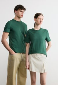 Άνδρας και γυναίκα φορούν ταιριαστά πράσινα μπλουζάκια, ο άνδρας με μπεζ παντελόνι και τα χέρια στις τσέπες, η γυναίκα με ανοιχτό μπεζ φούστα.