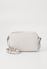 Ljust beige läder crossbody-väska med präglad "cK"-logotyp, dragkedjestängning och justerbar rem. Slät textur, rektangulär form.