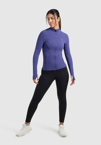 Lilla zip-up jakke lavet af strækbart materiale, med lange ærmer med tommelhul og sidelommer, parret med sorte leggings og hvide sneakers.