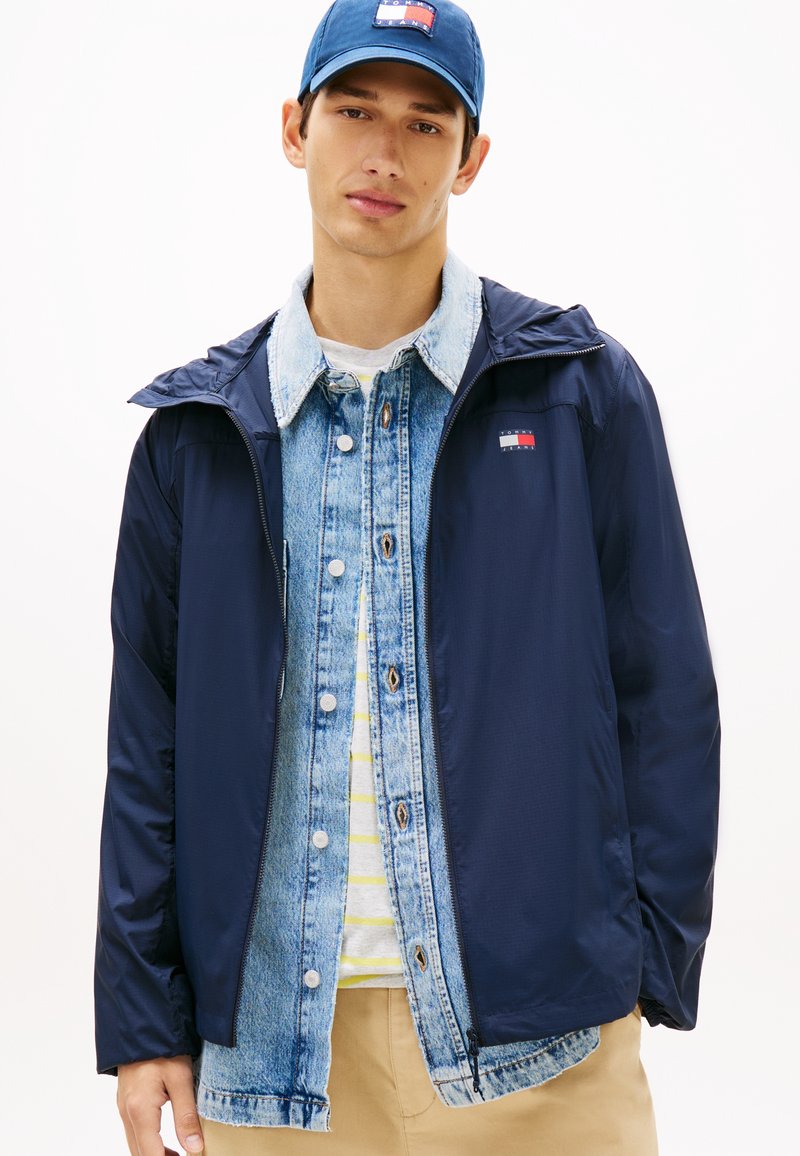 Νεαρός άνδρας που φοράει στρωματοποιημένο μπλε σκουρόχρωμο μπουφάν, μπλε τζιν πουκάμισο, ριγέ λευκό και κίτρινο T-shirt, μπεζ παντελόνι και μπλε σκουρόχρωμο καπέλο μπέιζμπολ.