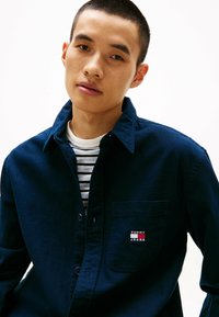 Camicia button-up blu navy in tessuto di cotone, con una tasca sul petto che presenta un logo "TOMMY JEANS" in contrasto. Indossata sopra una maglietta a righe bianca e blu navy.
