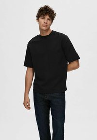 Tricou negru cu mânecă scurtă, din bumbac, cu guler rotund, purtat cu o pereche de blugi negri, stând într-un fond simplu.