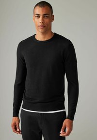 Sort langærmet sweater i blødt strikmateriale, rund halsudskæring og tætsiddende design; parret med sorte bukser og en hvid undertrøje.