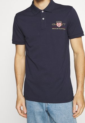 Mężczyzna noszący granatową koszulkę polo z logo GANT 1949 American Sportswear, w połączeniu z jasnoniebieskimi jeansami, stojący na białym tle.