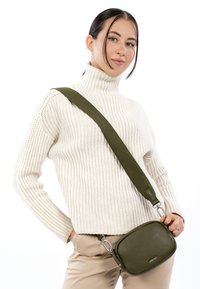 Alyvuogių žalia odinė rankinė su tekstūruota paviršiaus danga, reguliuojama drobinė petneša ir sidabro spalvos detalės. Dėvima kryžminiu būdu ant šviesaus megztinio.