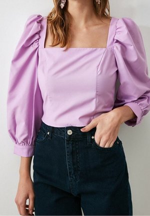 Kobieta w liliowej bluzce z bufkami i kwadratowym dekoltem, zestawiona z ciemnoniebieskimi jeansami z wysokim stanem, ręka w kieszeni.