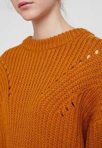 Oranger gestrickter Pullover mit strukturiertem Rippmuster, der offene Maschendetails nahe der Schulter und einen breiten Rippkragen aufweist.