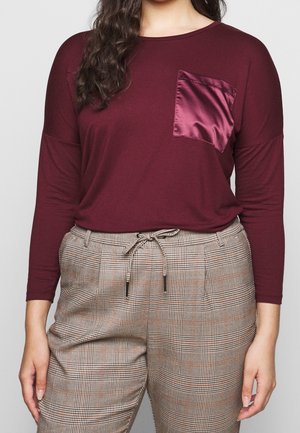 Burgundowa bluzka z długim rękawem z satynową kieszonką na piersi, zestawiona z wężowymi beżowo-brązowymi spodniami z troczkiem w talii.
