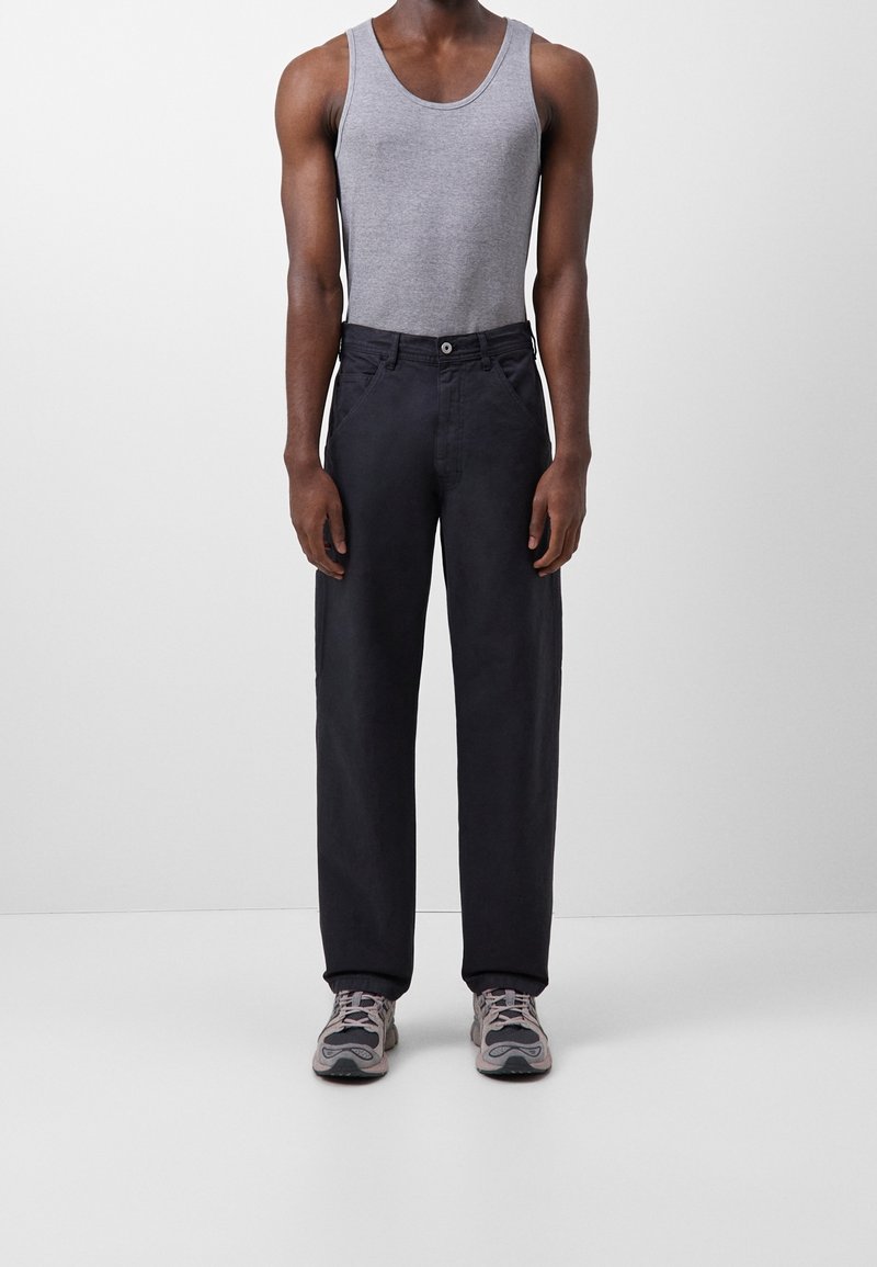 Homme portant un débardeur gris, un pantalon large foncé et des baskets grises, debout devant un fond gris clair uni.