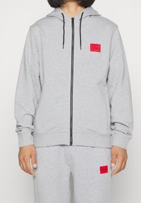 Sudadera gris con cremallera hecha de tela suave, con capucha y cordones negros. Presenta un logo cuadrado rojo en el pecho y puños y dobladillo acanalados.