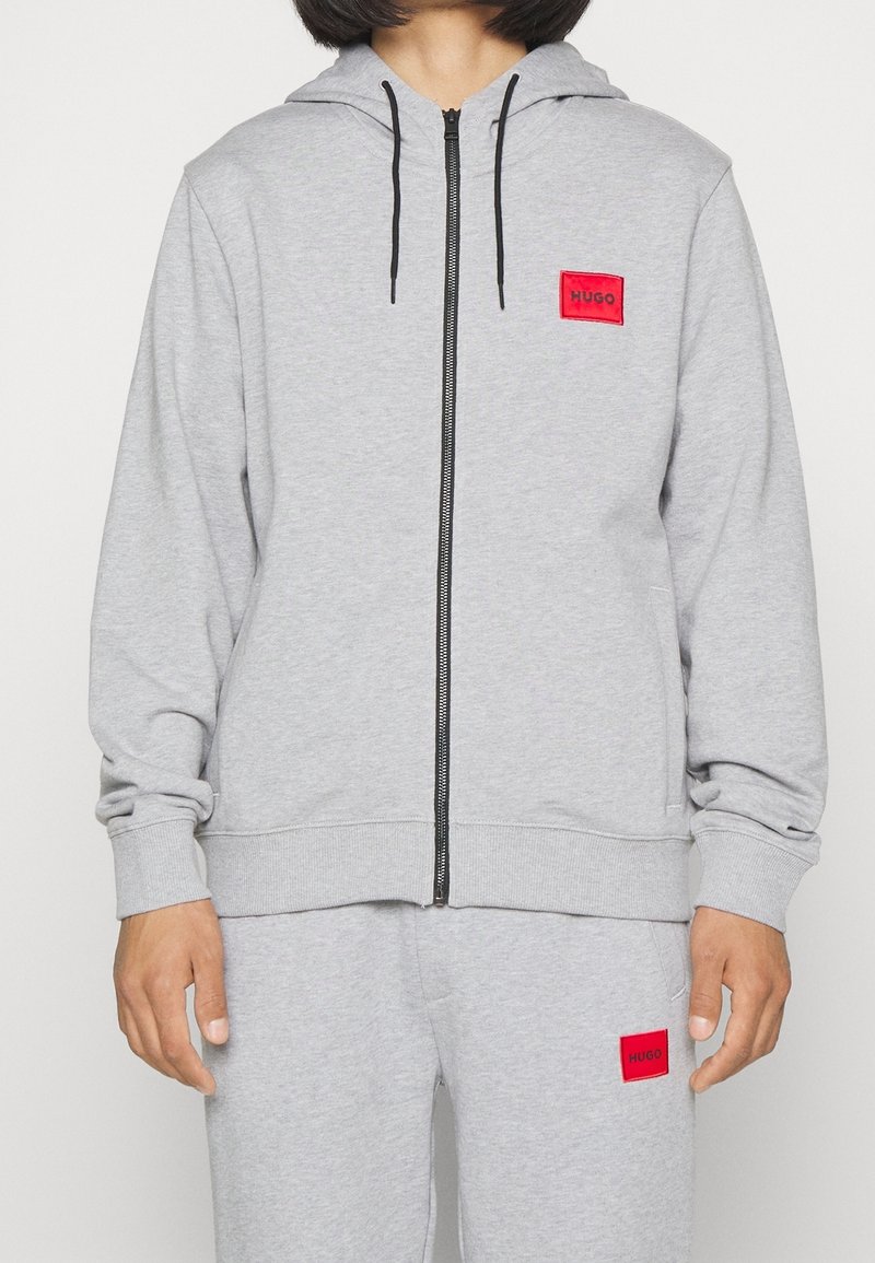Sudadera gris con cremallera hecha de tela suave, con capucha y cordones negros. Presenta un logo cuadrado rojo en el pecho y puños y dobladillo acanalados.