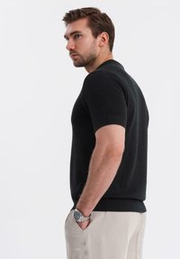 Maglione a maniche corte nero in maglia con colletto e polsini a coste, caratterizzato da un motivo texturizzato. Indossato con pantaloni chiari.