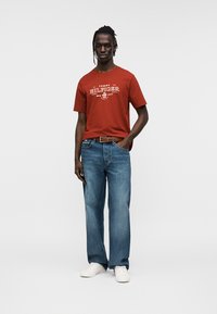 Κόκκινο βαμβακερό T-shirt με λευκό γραφικό κείμενο, σε συνδυασμό με μπλε τζιν παντελόνι. Λευκά αθλητικά παπούτσια ολοκληρώνουν το καθημερινό ντύσιμο. Το μοντέλο στέκεται με αυτοπεποίθηση.