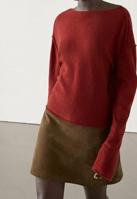 Pull rouge en mélange de laine avec un large décolleté et des manches amples, associé à une mini-jupe en daim marron, créant un look superposé.