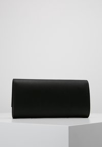 Clutch de cuero sintético negro con forma cilíndrica, textura lisa y sin herrajes ni adornos visibles, colocado sobre una superficie blanca.