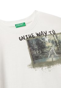Bílý tričko s grafikou lesní cesty a textem "NA CESTĚ K" nad ním, zelená značka "United Colors of Benetton" uvnitř límce.