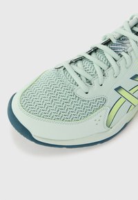 Botte de sport vert clair avec tige en mesh, bout rond et semelle extérieure bleue. Présente des lacets et des bandes d'accent jaune sur le côté.