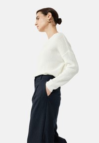 Crème ribgebreide trui met lange mouwen, gecombineerd met donker gestreepte high-waisted broek. De stof lijkt zacht en gestructureerd.